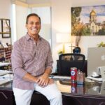 Holy Trinity principal Anthony DiLeva sits on a desk in an office smiling, wearing a checkered button-down shirt.