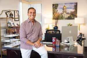 Holy Trinity principal Anthony DiLeva sits on a desk in an office smiling, wearing a checkered button-down shirt.